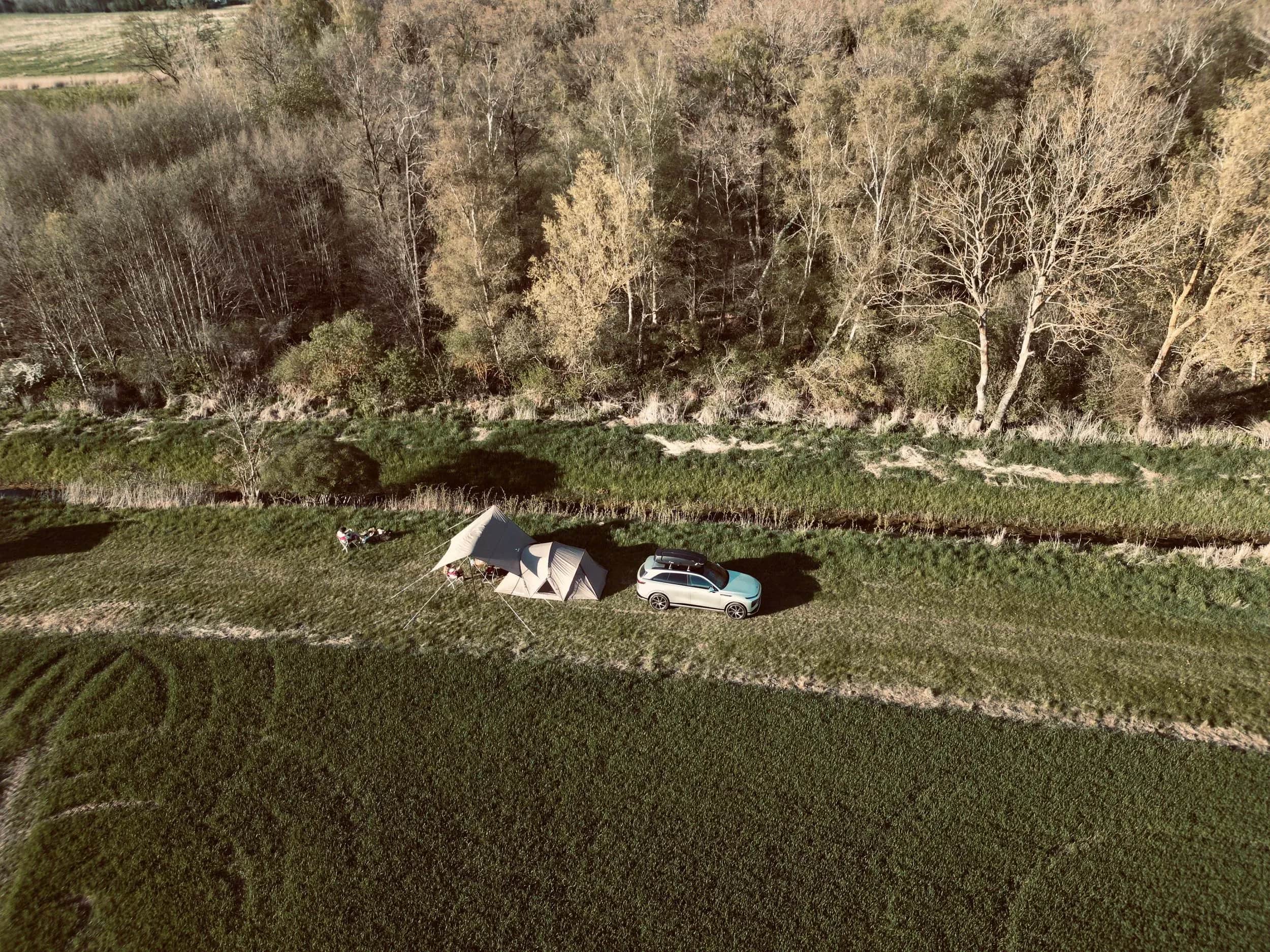 Luftfoto af telte og en bil ved en å langs en mark.