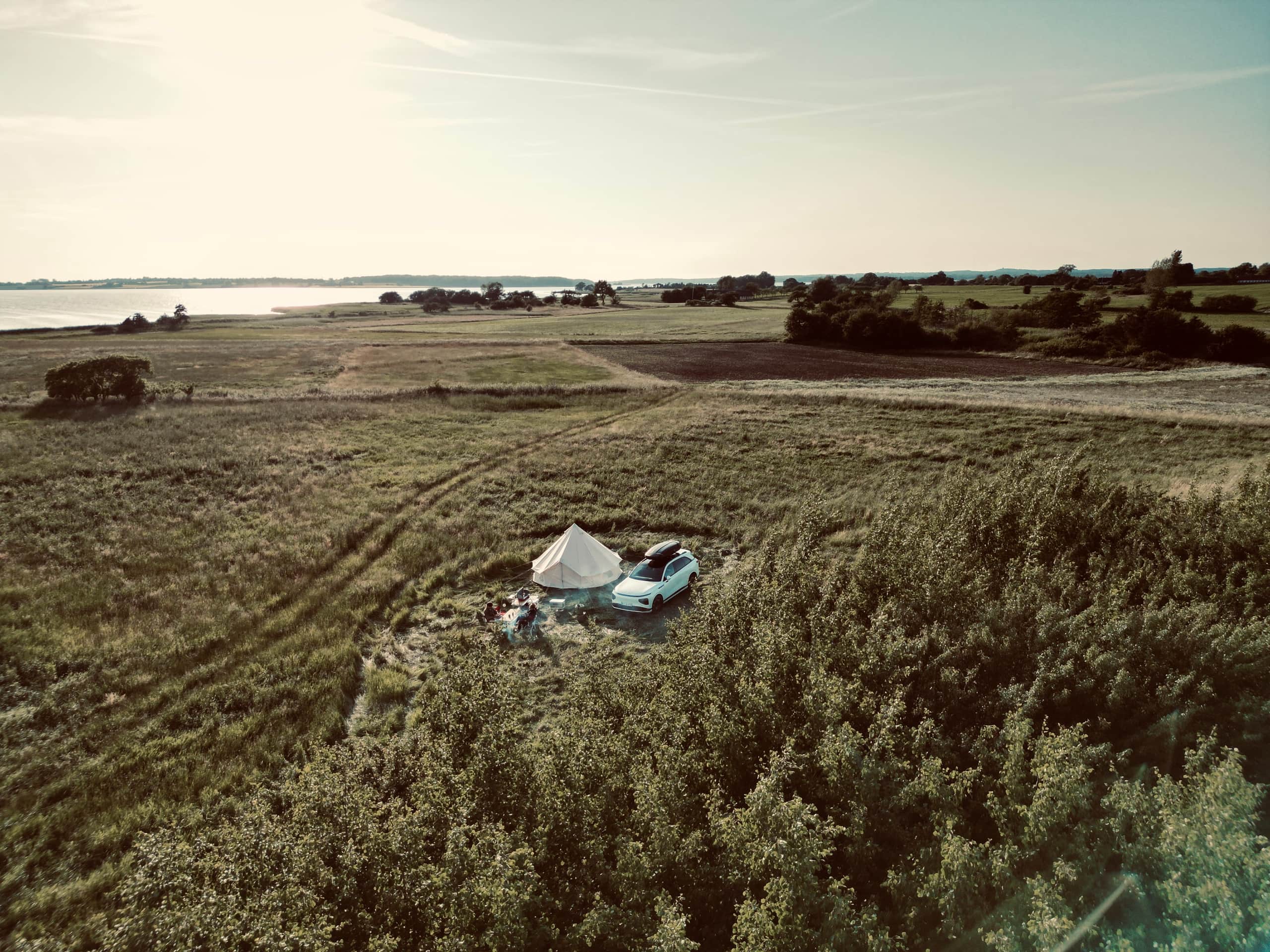 Luftaufnahme eines beigen Zelts und eines weißen Autos auf einem Naturzeltplatz zwischen Feldern mit Blick aufs Wasser.